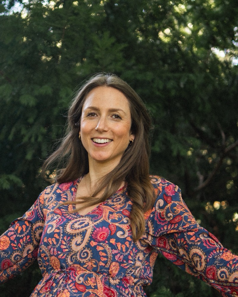 Jasmin Gaschler lachend mit ausgebreiteten Armen vor Bäumen, im gemusterten Kleid